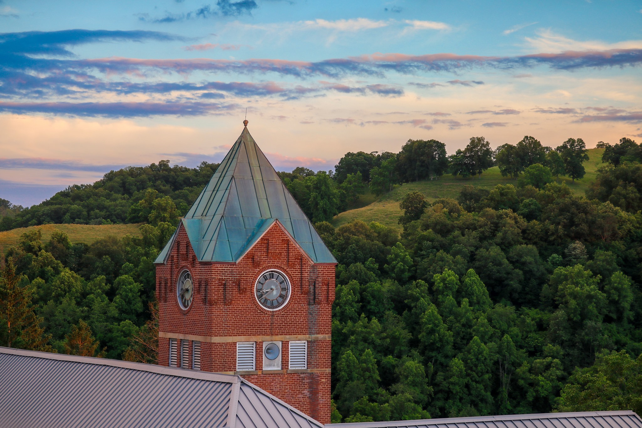 The GSC Clock Tower stands tall in front of rising green hills speckled with trees and meadows. It is dusk, and the sky shifts from purple, to pale pink, to light blue.