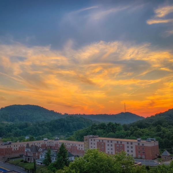 A wide arial shot of campus at sunset. The campus is tucked among the hills, which have faded to dark greens and blues. The sky is awash with blazing oranges and reds.