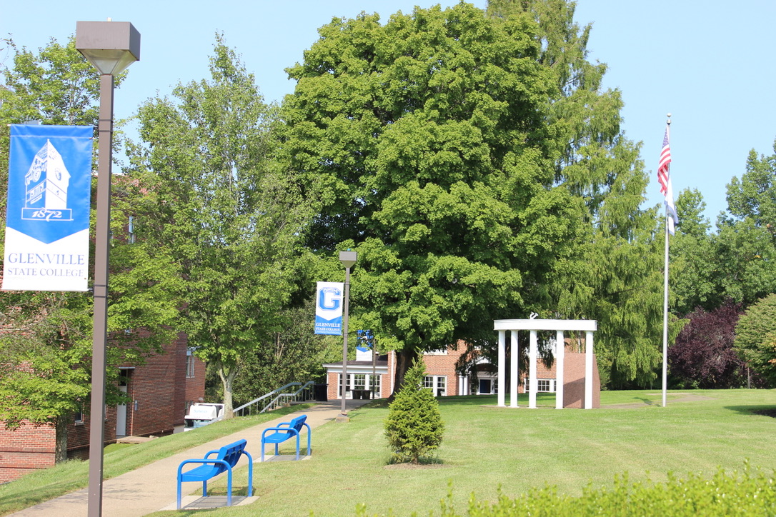 A photo of Glenville State College in summer, with several blue banners hanging from light poles and bright blue benches lining a cement pathway. Mature trees surround the area.