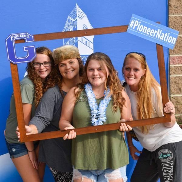 Four female students pose for a photo while holding an oversize wooden frame, decorated with messages like "Pioneer Nation" and "GSC Pioneers." Behind them is a large blue backdrop with the Glenville State logo on it.