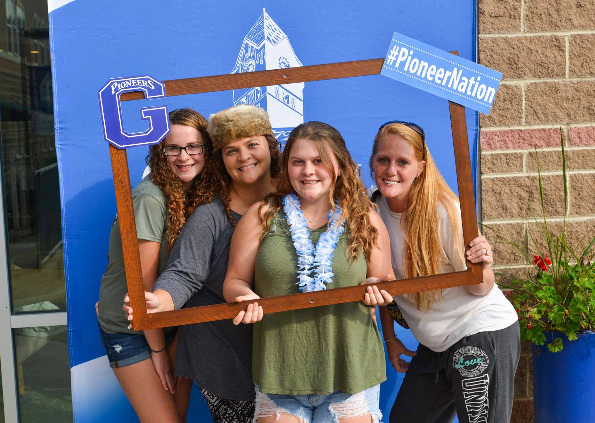 Four female students pose for a photo while holding an oversize wooden frame, decorated with messages like "Pioneer Nation" and "GSC Pioneers." Behind them is a large blue backdrop with the Glenville State logo on it.