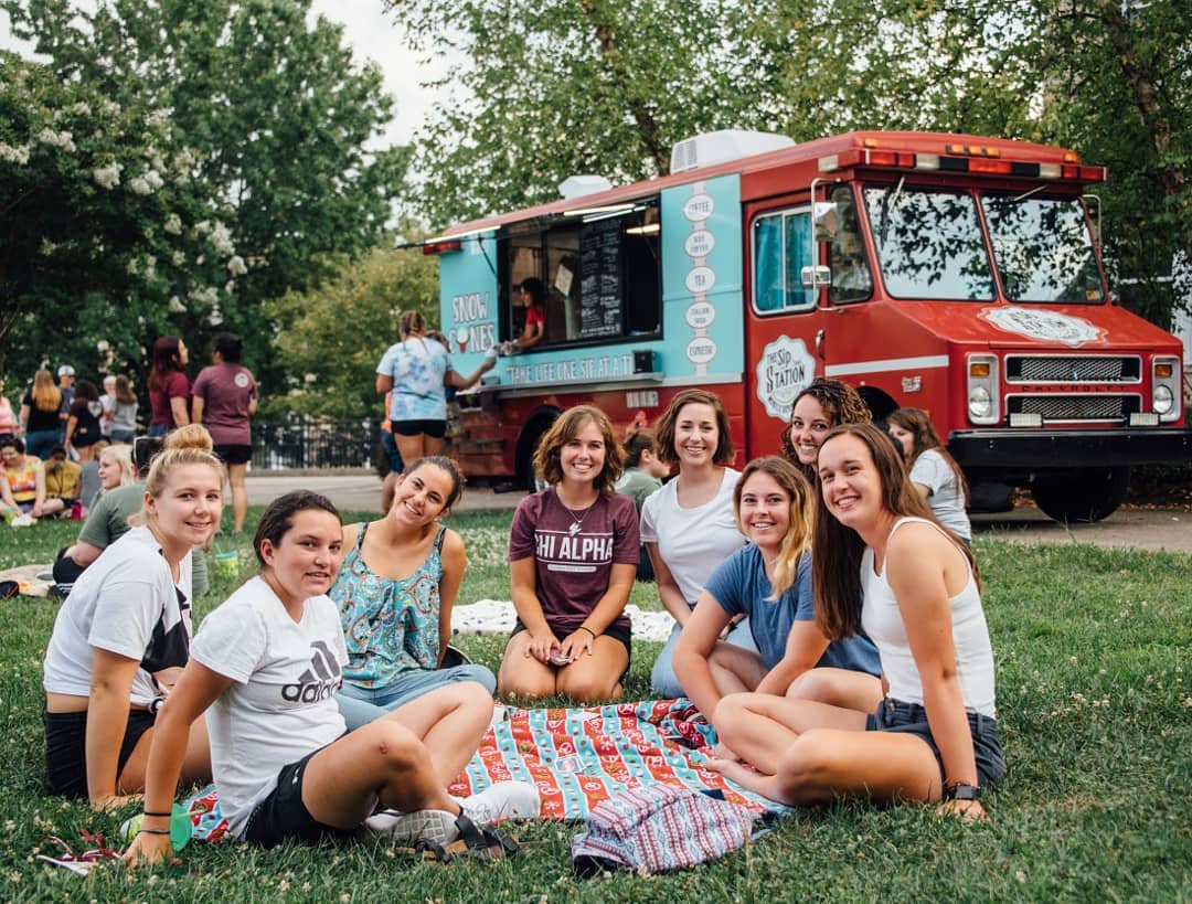 A group of students sit on a picnic blanket on the campus quad. In the background, you can see a colorful snow cone truck.