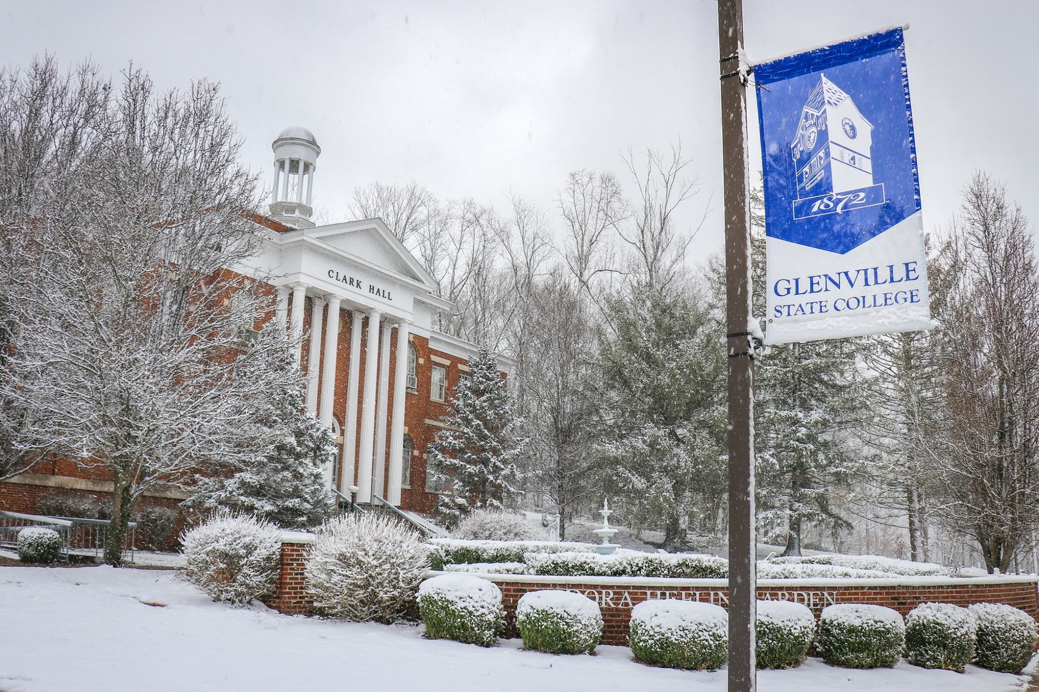 Heavy snow falls on the garden in front of the stately Clark Hall. In the foreground, a brilliant blue Glenville State College banner hangs from a lamp post. Trees, bushes and the ground are covered with several inches of snow.