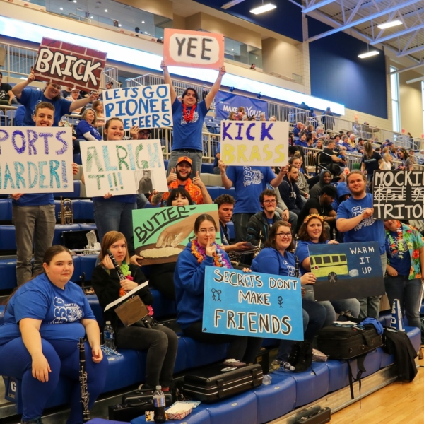 Students line the stands of the GSC basketball arena. They are holding homemade signs with messages like ""Kick Brass," "Let's Go Pioneers," and "Sports Harder!"