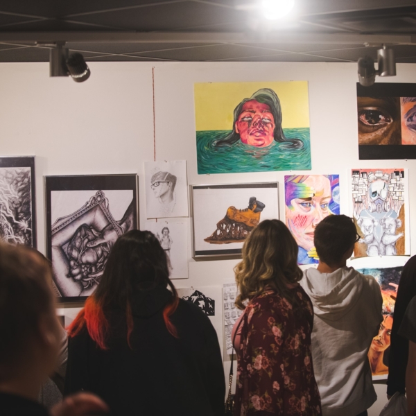 A group of spectators examine student artwork, hanging on a gallery wall