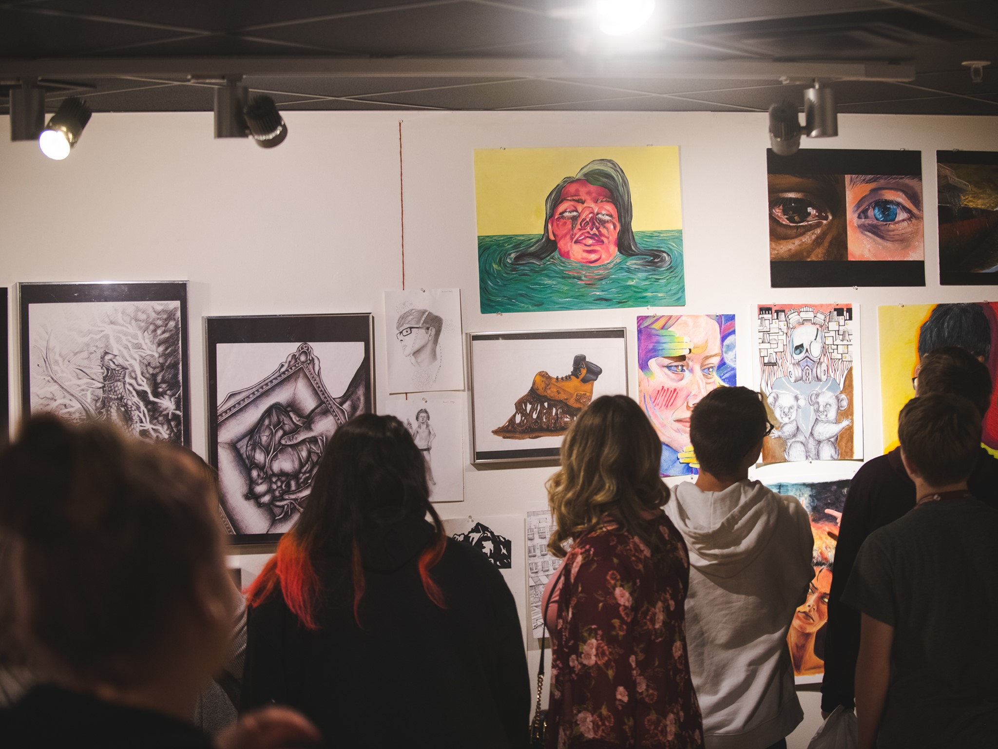 A group of spectators examine student artwork, hanging on a gallery wall