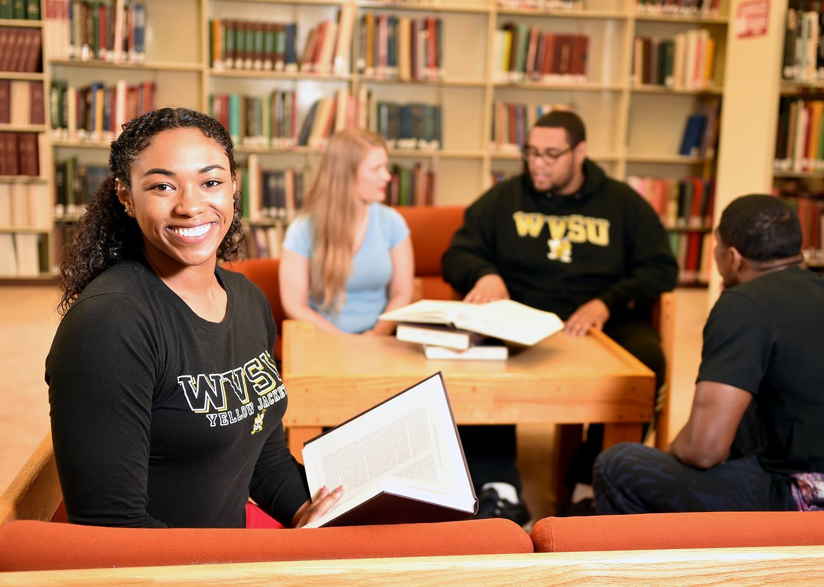 In the library, a student smiles while reading a book in a WVSU shirt. Three students study in the background.