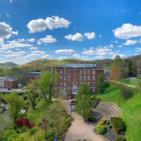 A wide arial shot of campus, which is tucked amongst the West Virginia hills. It's a sunny day with a brilliant blue sky and bright fluffy clouds. Trees are just beginning to show new leaves and the grass is an almost-neon green from new growth. In the center of the picture stands Science Hall and the amphitheater.