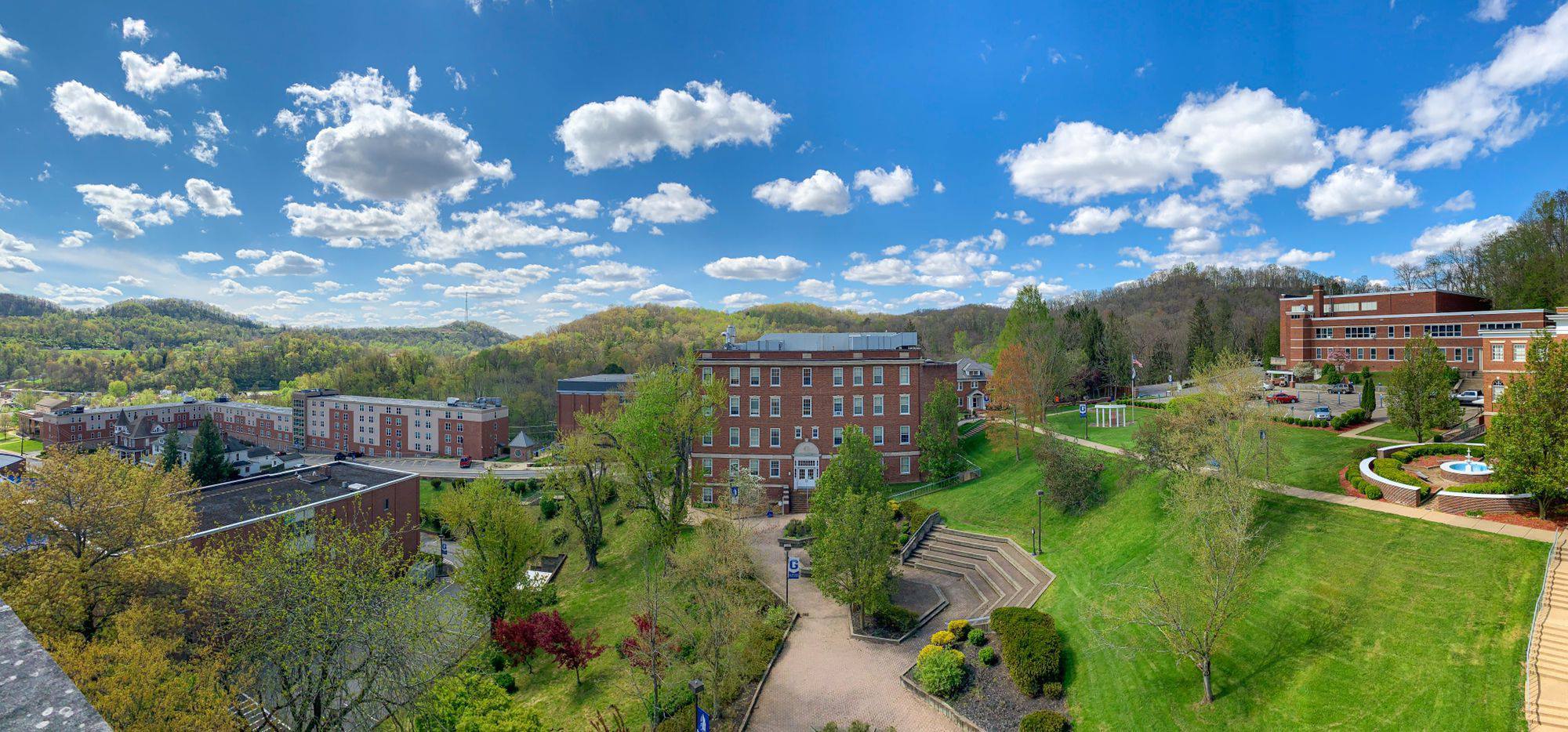 A wide arial shot of campus, which is tucked amongst the West Virginia hills. It's a sunny day with a brilliant blue sky and bright fluffy clouds. Trees are just beginning to show new leaves and the grass is an almost-neon green from new growth. In the center of the picture stands Science Hall and the amphitheater.