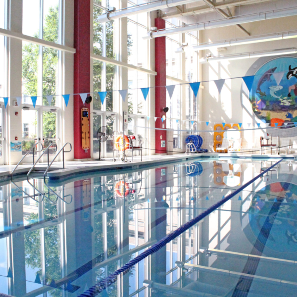 A pool with various swimming lanes is situated in a large room with floor to ceiling windows. A volleyball net hangs in the center of the pool.