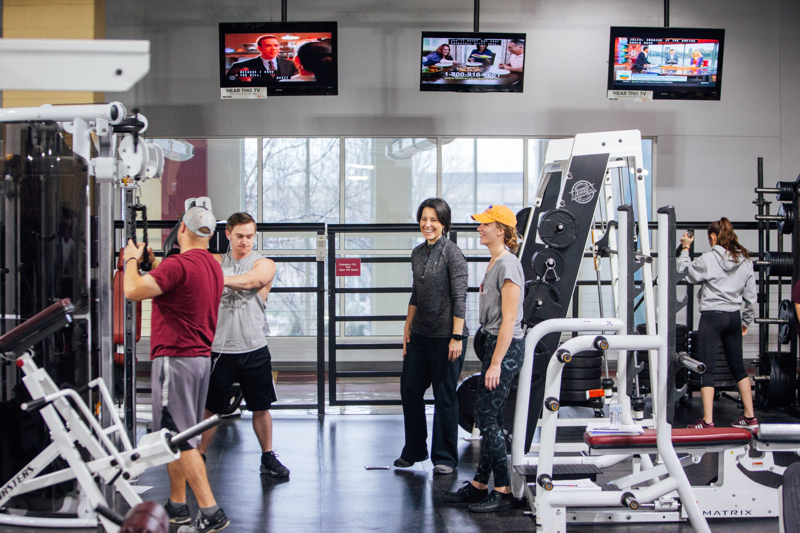 Students use a variety of weight training equipment in a modern-looking indoor gym.