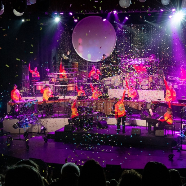 The GSC percussion ensemble performs on a stage illuminated with pink, purple, and blue lights. Confetti fills the air as the percussionists raise their drum sticks in a final salute to end the show.