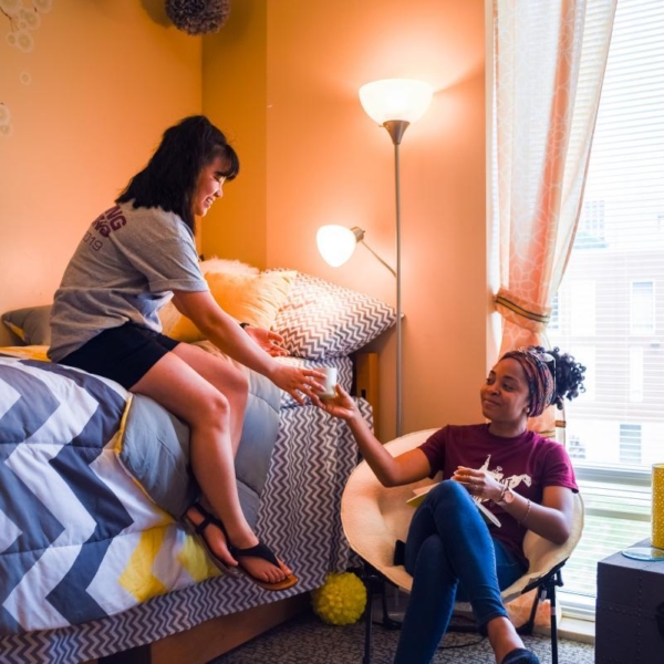 Two students sit in front of a large window in a residence hall room, which contains two twin beds, a large chair, flowers, and floral curtains