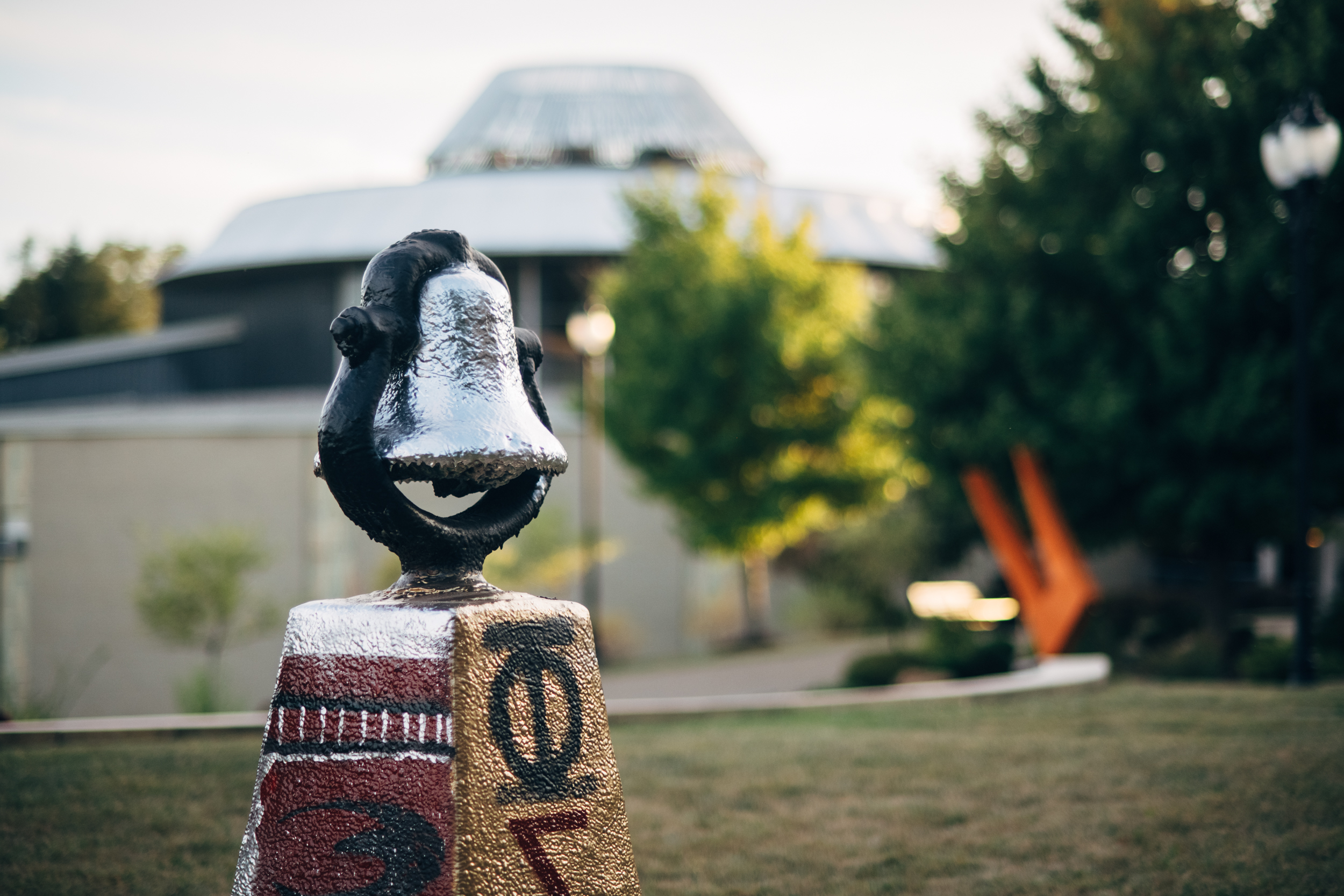 A close-up of the campus victory bell, which is painted silver and black. The base the bell sits on is painted maroon, gold, silver, and black, and features Greek letters. In the background, you can see the soft outlines of a bright orange abstract sculpture.